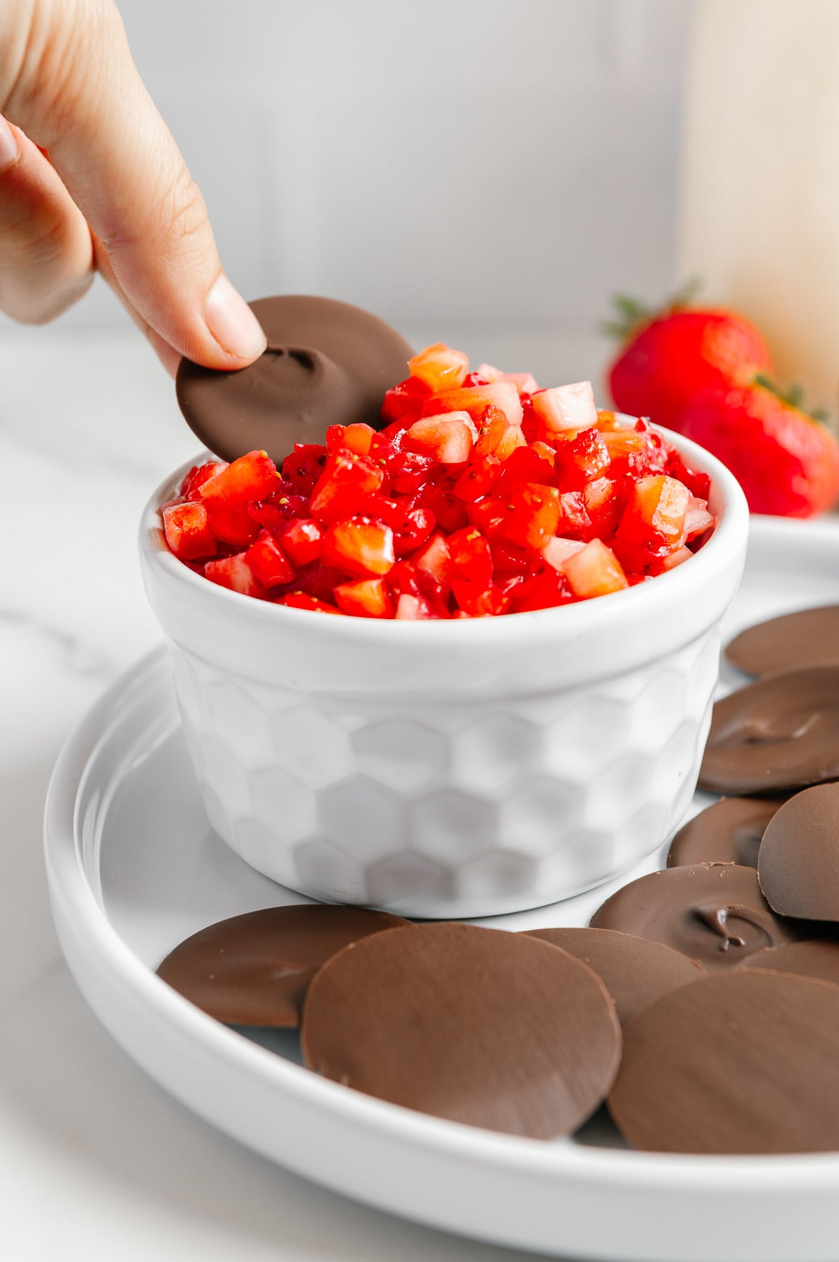 A hand dipping a chocolate disc into a small white bowl of strawberry "salsa."