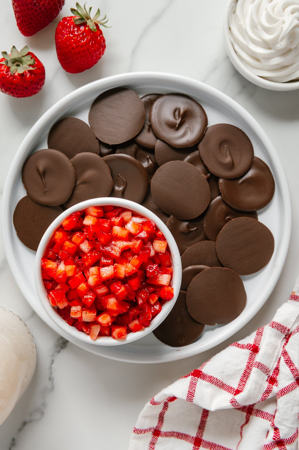 Strawberry "salsa" in a small white bowl on a white plate with chocolate "chips" next to fresh strawberries and coconut whipped cream on a white marble background.