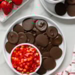 Chocolate discs and strawberry salsa on white plates alongside fresh strawberries and a white and red checkered towel with a white marble background.