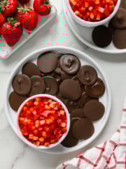 Chocolate discs and strawberry salsa on white plates alongside fresh strawberries and a white and red checkered towel with a white marble background.