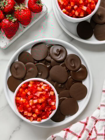 Chocolate discs and strawberry salsa on white plates alongside fresh strawberries and a white and red checkered towel with a white marble background.
