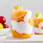 Apple cider slushies served with coconut cream and garnished with apple slices on a white block with a bowl of fresh apples in the background.