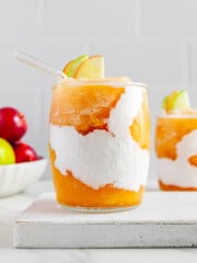 Apple cider slushies served with coconut cream and garnished with apple slices on a white block with a bowl of fresh apples in the background.