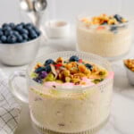 Two glass mugs of overnight oats next to a bowl of fresh blueberries with a white marble background.