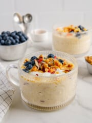 Two glass mugs of overnight oats next to a bowl of fresh blueberries with a white marble background.
