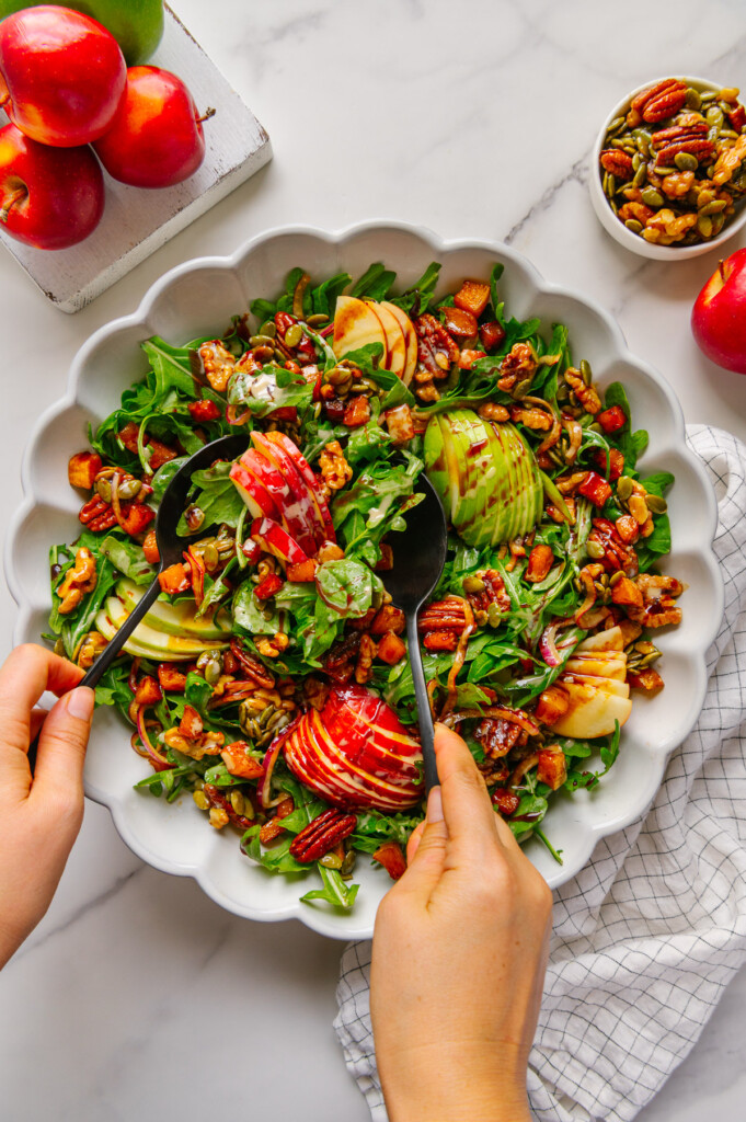 Hands using spoons to toss a vegan fall harvest salad in a white bowl.