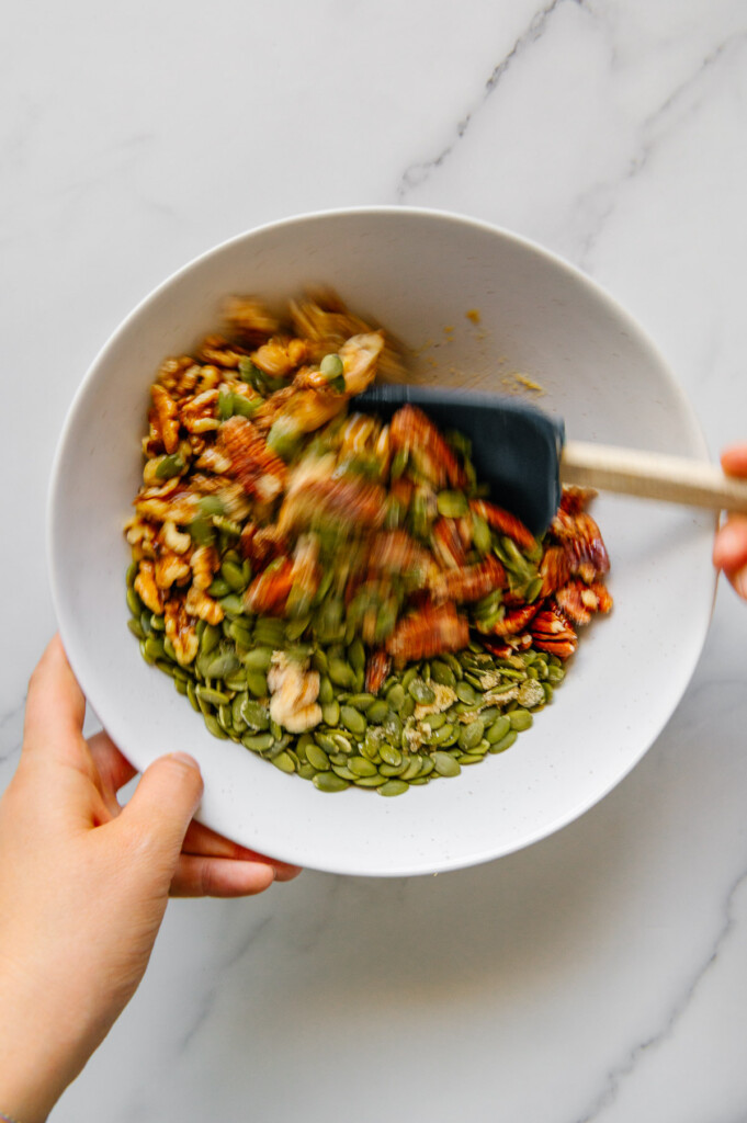Hands using a spatula to mix nuts and seeds in a white bowl.