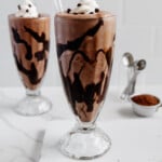 Vegan chocolate milkshakes in two tall glasses with a small measuring cup of cocoa powder in the background with a white marble background.