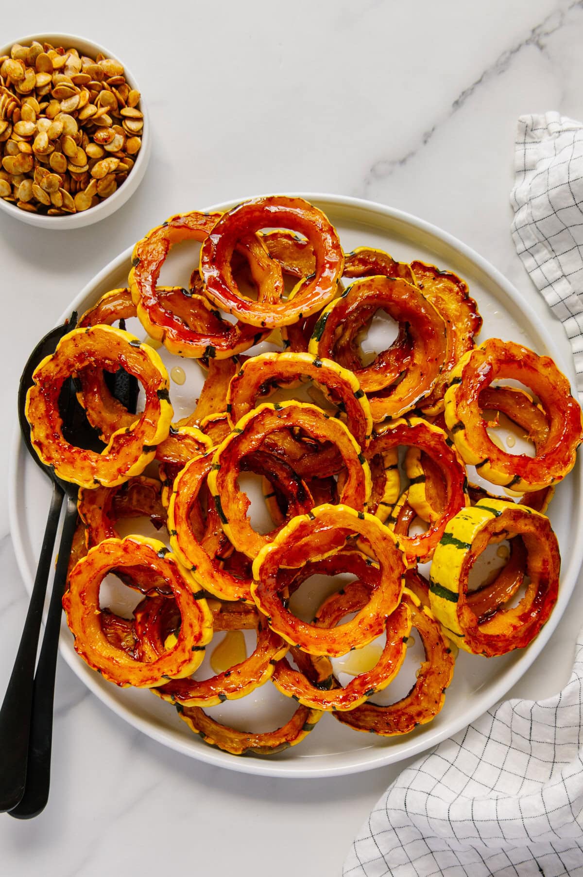 Roasted delicata squash on a white plate next to a bowl of delicata squash seeds with a white marble background.