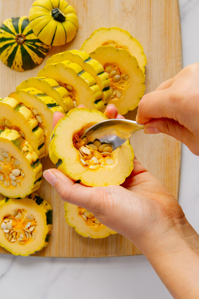 Hands using a spoon to scoop out seeds from a delicata squash ring above more squash rings on a wooden cutting board.