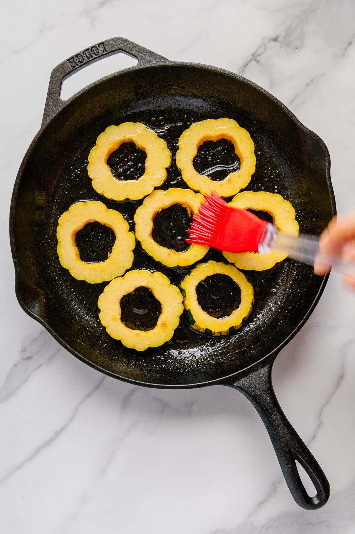 A silicone brush brushing oil onto sliced rings of delicata squash in a cast iron skillet.