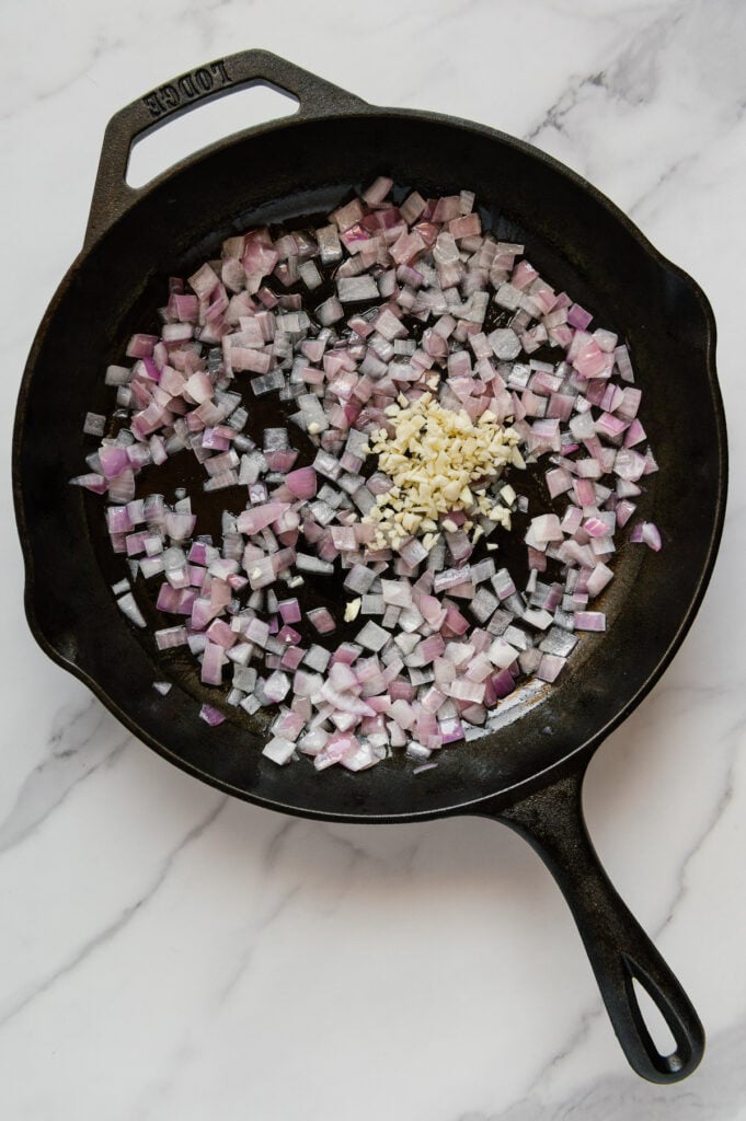 Diced red onions and fresh garlic in a cast iron skillet with a white marble background.