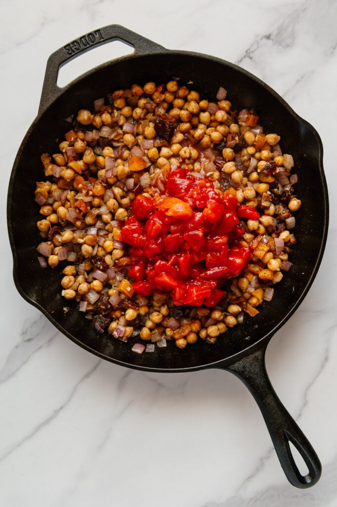 Diced tomatoes over Moroccan chickpea stew ingredients in a cast iron skillet with a white marble background.