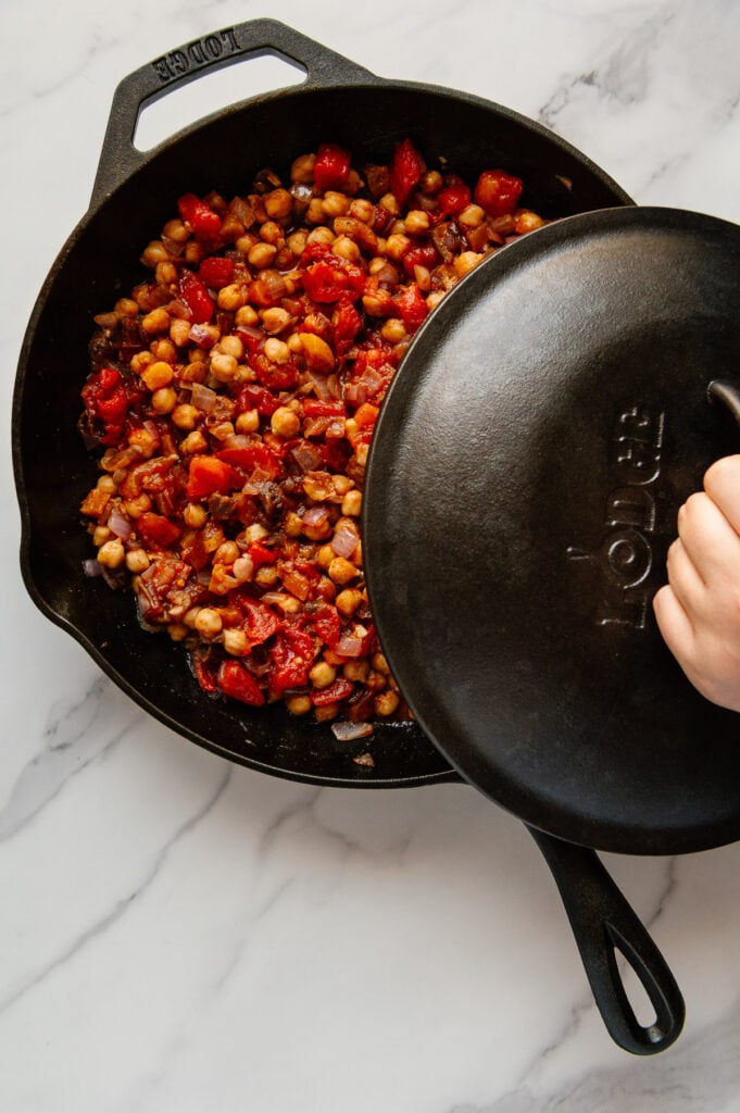 A hand putting a cover over a skillet of Moroccan chickpeas with a white marble background.