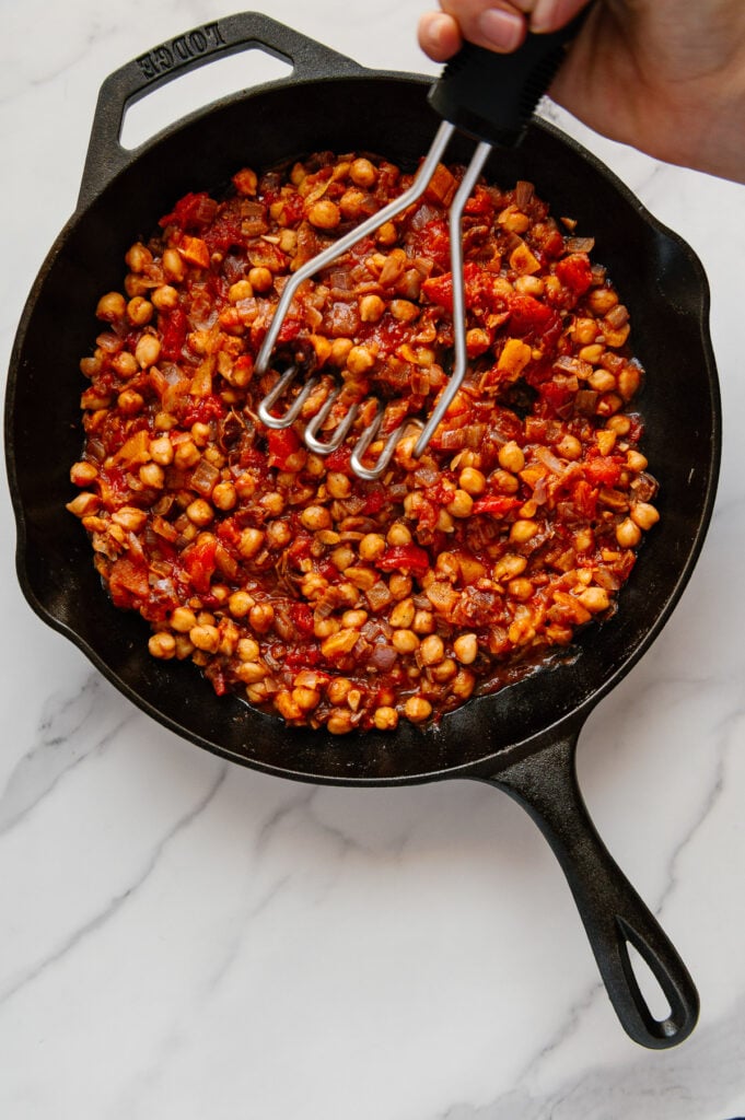 A hand using a potato masher to mash chickpeas in a cast iron skillet of Moroccan chickpea stew with a white marble background.