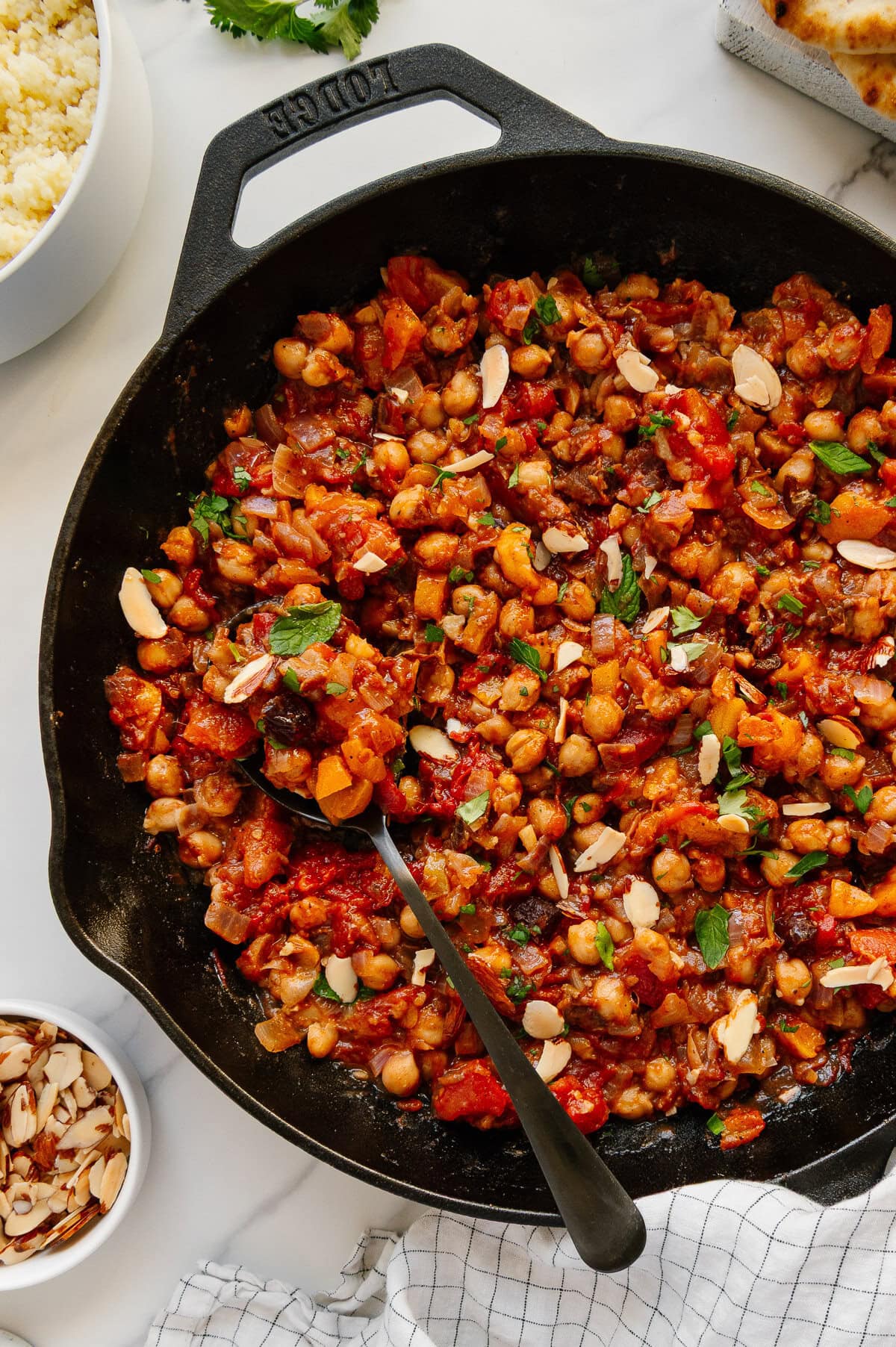 Moroccan chickpeas garnished with toasted sliced almonds and fresh herbs in a cast iron skillet with a white marble background.