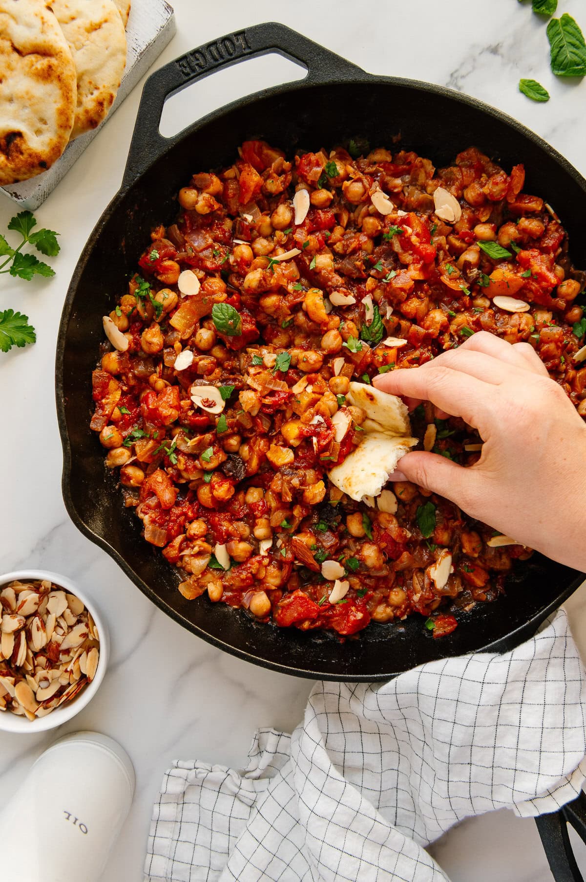 A hand dipping a soft pita into a skillet of Moroccan chickpeas with a white marble background.