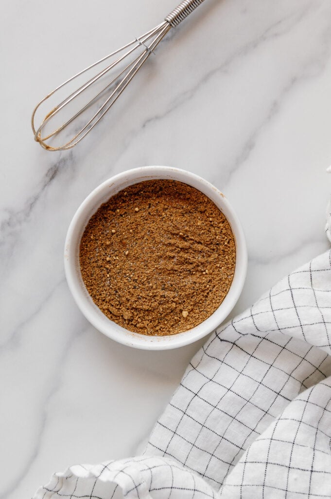 Ras el hanout spice blend in a small white bowl next to a whisk and white dish cloth with a white marble background.