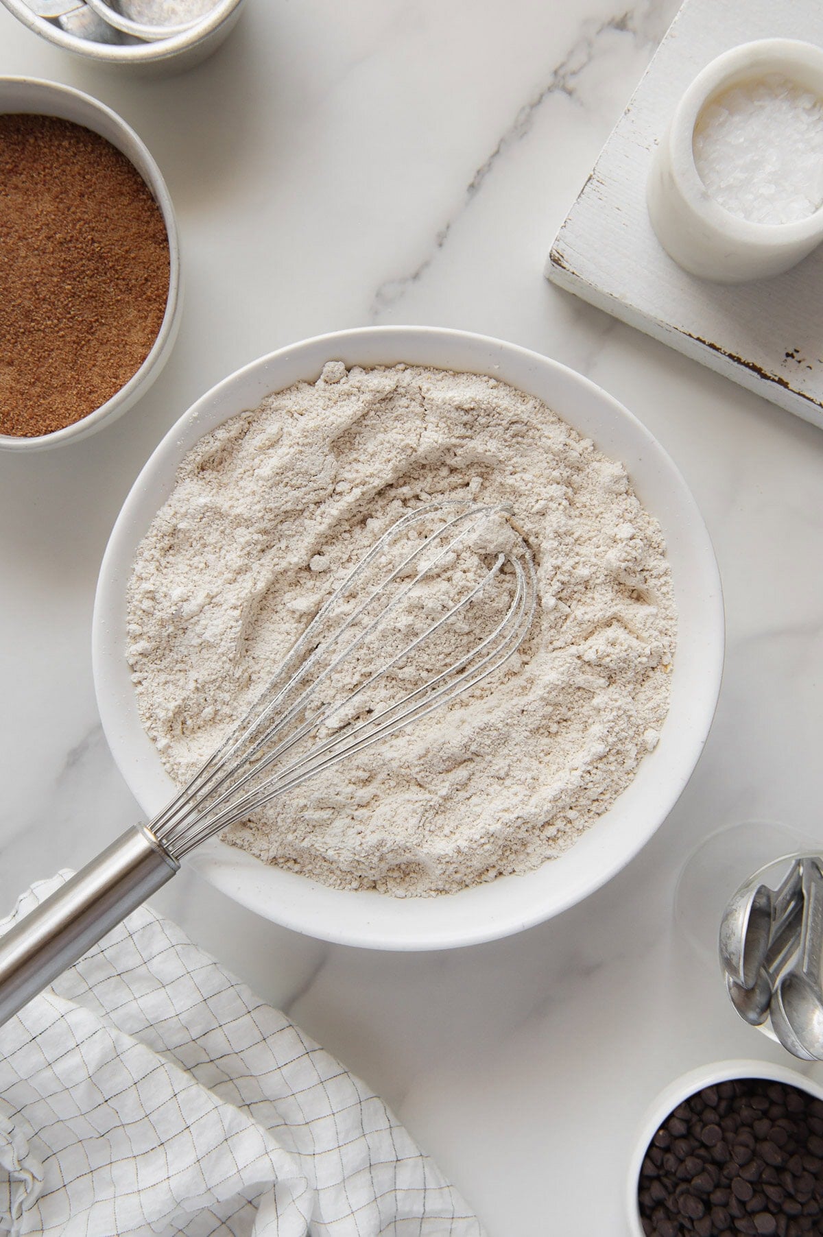 A whisk in a white bowl full of oat flour with a white marble background.