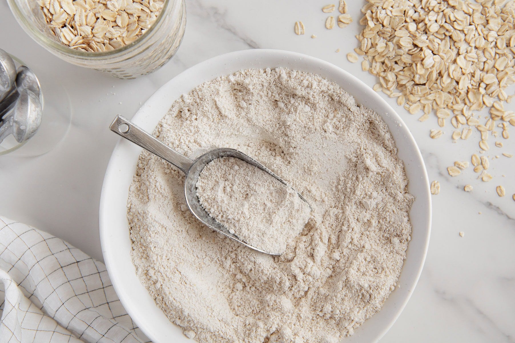 Oat flour in a bowl with a scooper next to oats with a white marble background.
