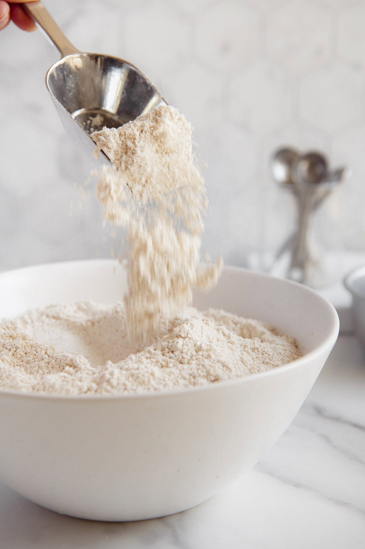 Oat flour being sprinkled from a scoop into a white bowl.