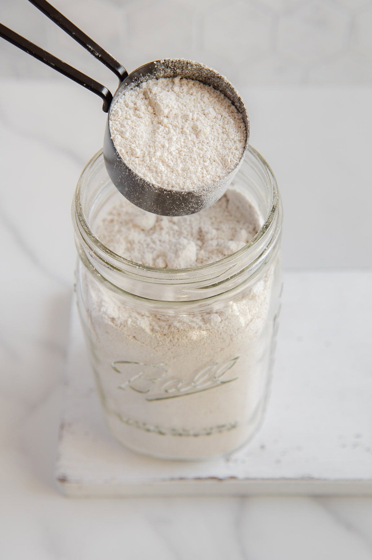 A measuring cup of oat flour held above a glass jar of oat flour with a white backround.