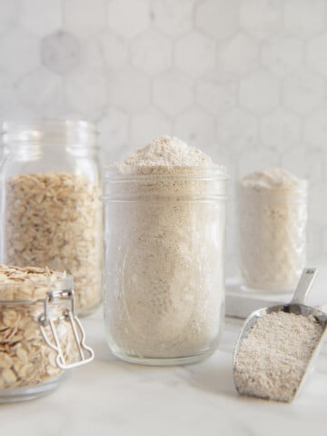 Glass jars filled with oats and oat flour next to a coop of oat flour with a white background.