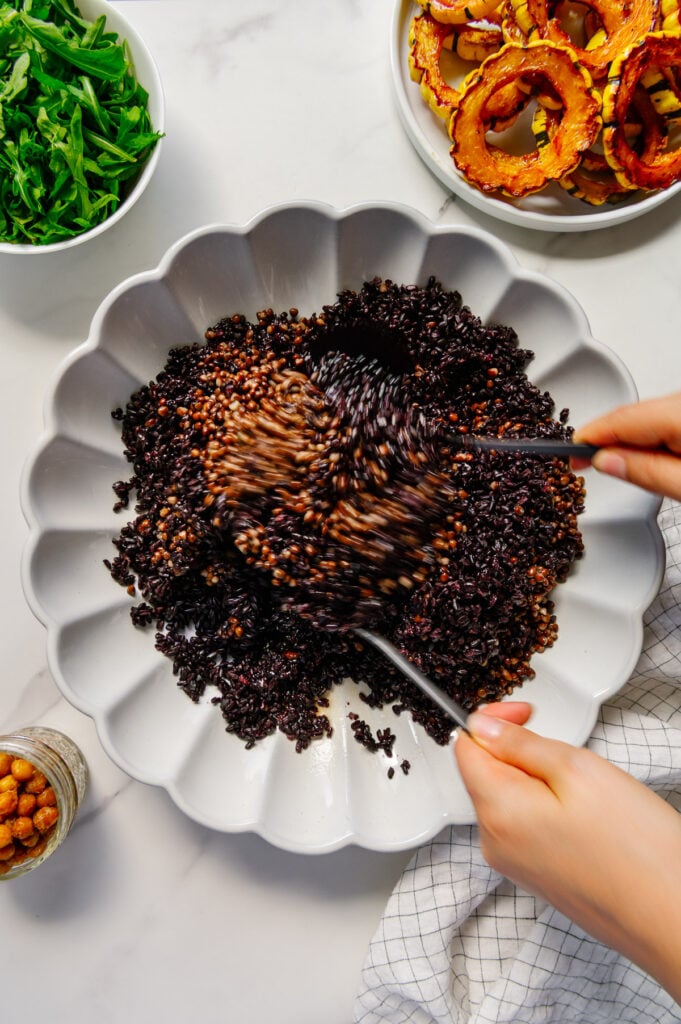 Using two spoons to mix black lentils and forbidden rice with dressing in a white serving plate.