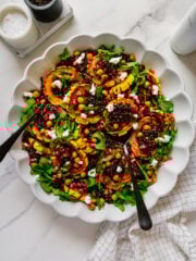 Maple roasted delicata squash rings and crispy chickpeas with lentils and forbidden rice topped with arugula, pomegranate seeds and vegan goat cheese on a white serving platter.