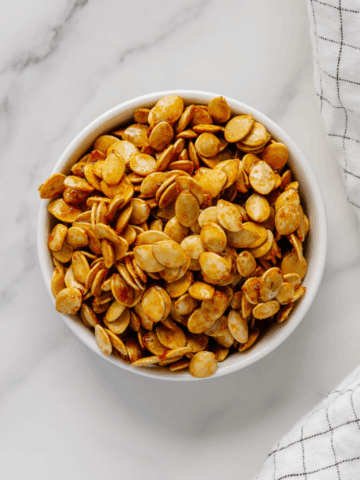 Roasted winter squash seeds in a white bowl with a white marble background.