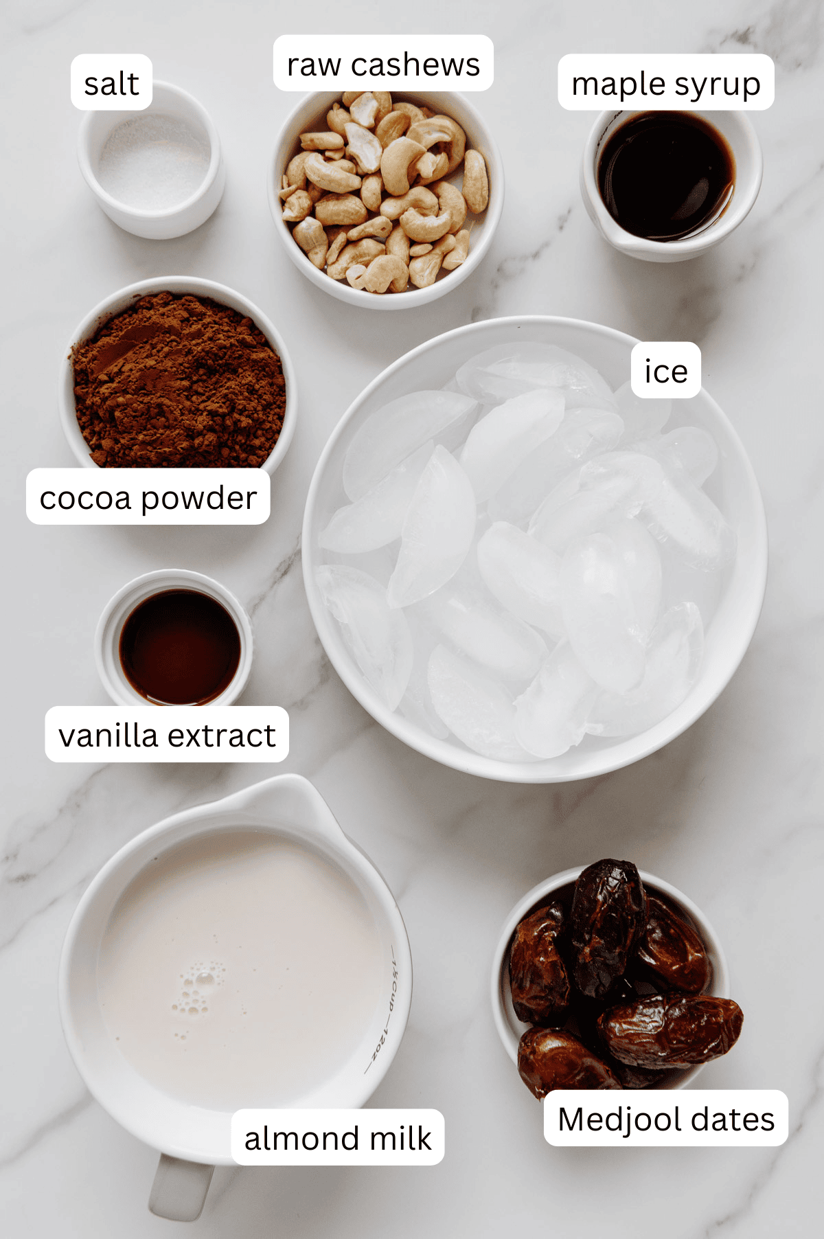 Vegan chocolate milkshake ingredients in separate bowls with a white marble background.