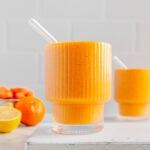 Two carrot smoothies in glasses with glass straws next to a bowl of clementines and a sliced lemon.