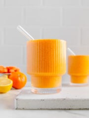 Two carrot smoothies in glasses with glass straws next to a bowl of clementines and a sliced lemon.