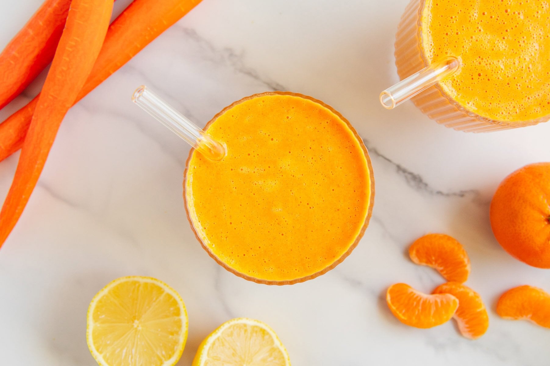 Overhead angle of a carrot smoothie in a glass with sliced carrot, lemon and clementines with a white marble background.