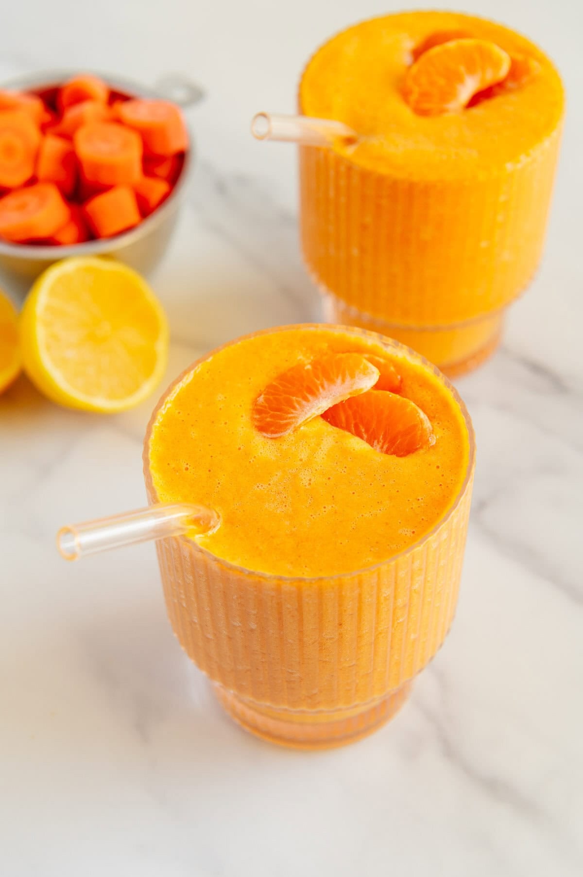 Two carrot smoothies in glasses garnished with clementines next to a bowl of sliced carrots and lemon slices with a white marble background.