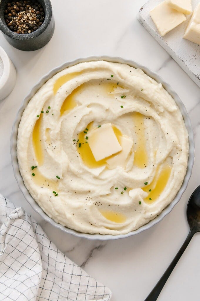 Mashed potatoes in a white bowl topped with butter and chives with a white marble background.