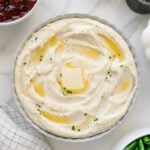 Mashed potatoes in a white bowl topped with butter and chives with a white marble background.