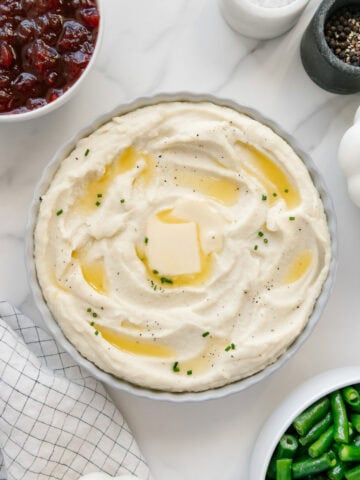Mashed potatoes in a white bowl topped with butter and chives with a white marble background.