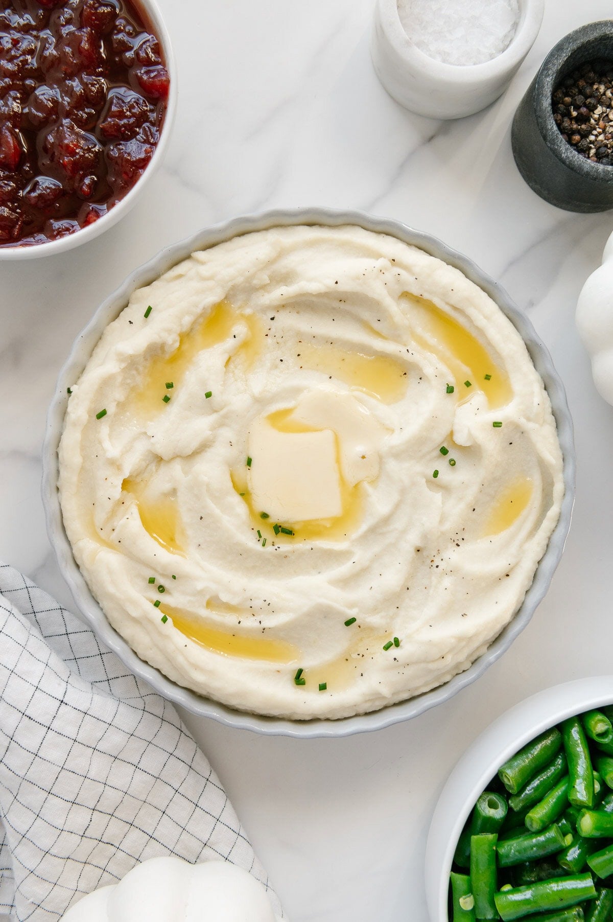 Mashed potatoes in a white bowl topped with butter and chives with a white marble background.