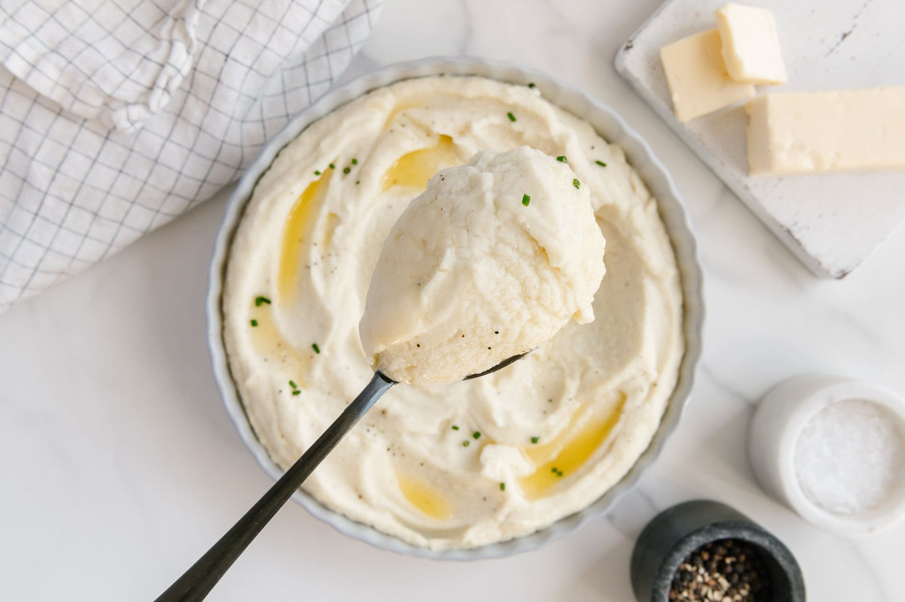A spoonful of mashed potatoes held over a white bowl of mashed potatoes next to a stick of vegan butter on a white platter and salt and pepper.