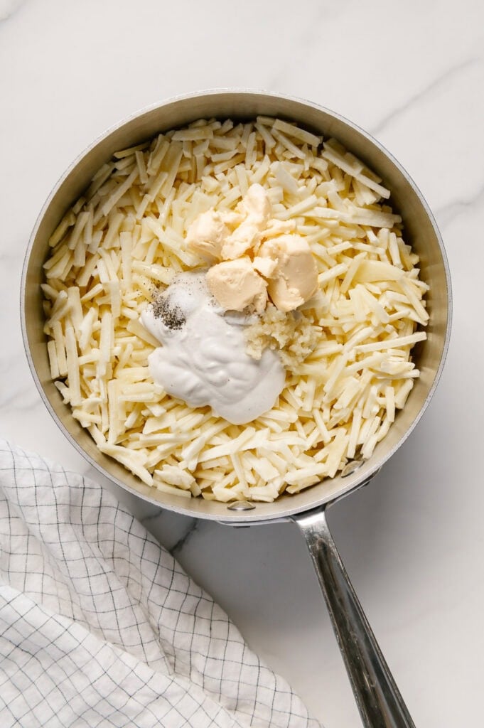 Frozen shredded potatoes, vegan butter, cashew cream and seasonings in a saucepan with a white marble background.