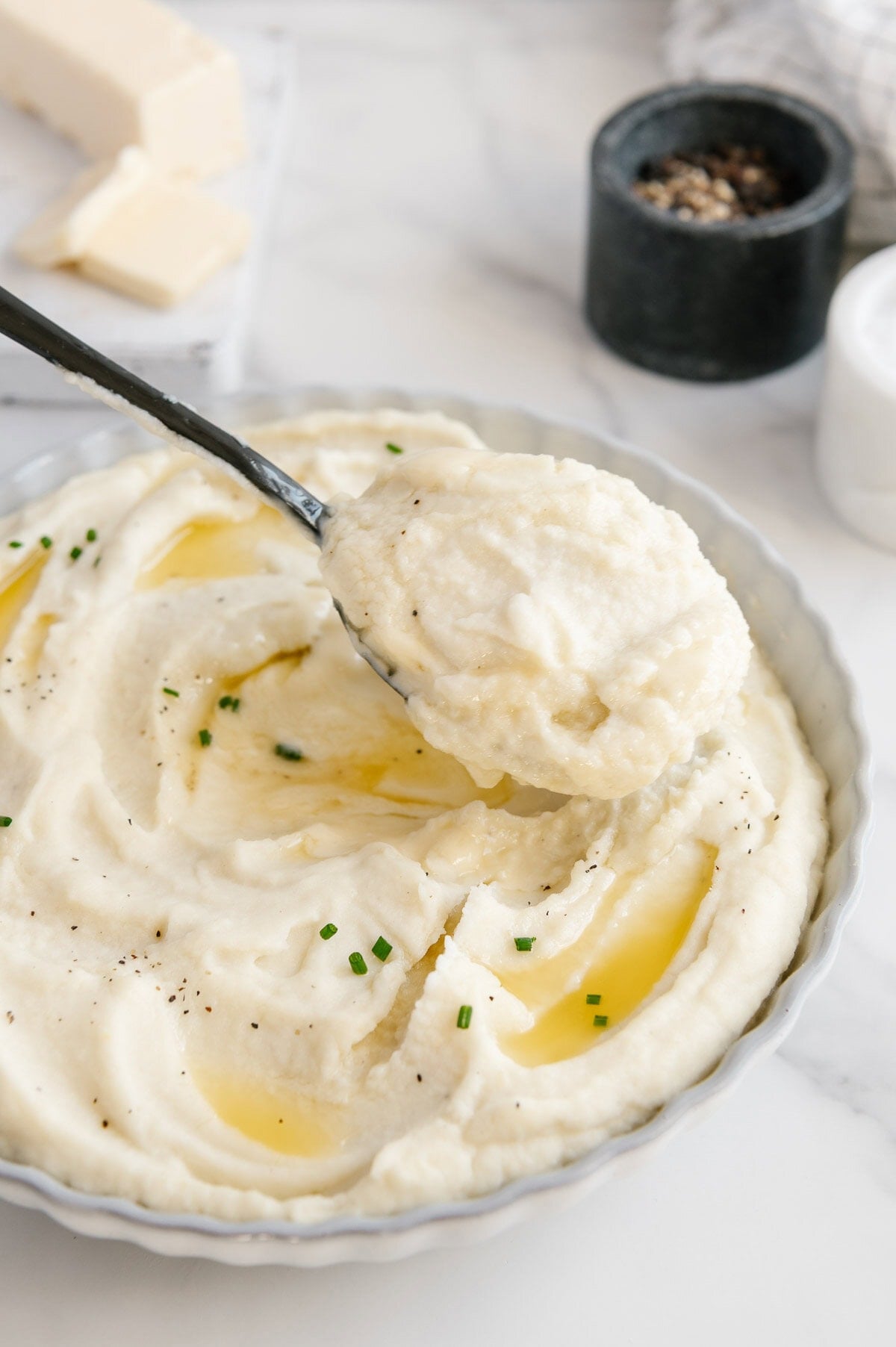 A spoonful of creamy mashed potatoes held above a white bowl of mashed potatoes topped with vegan butter and chives.