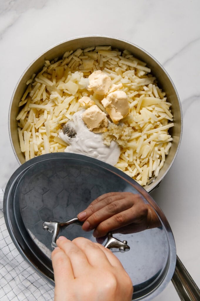 Frozen shredded potatoes, vegan butter, cashew cream and seasonings in a saucepan with a hand holding a lid with a white marble background.