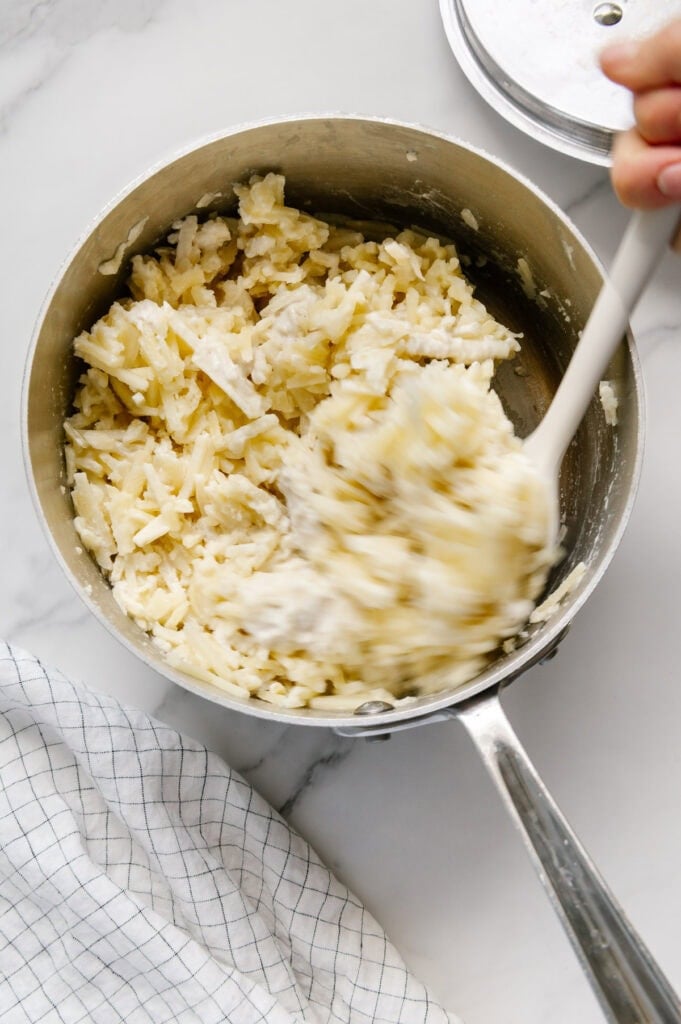 A hand using a white silicone spatula to mix potatoes in a saucepan.