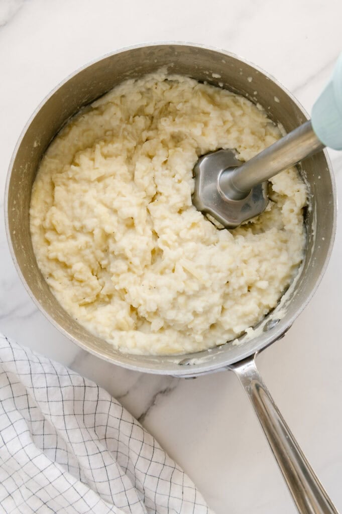 An immersion blender being used to pulse mashed potatoes in a saucepan.