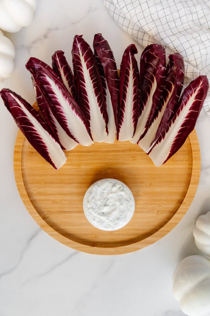 Circular wooden platter with a small bowl of vegan ranch at the bottom and endive leaves at the top.