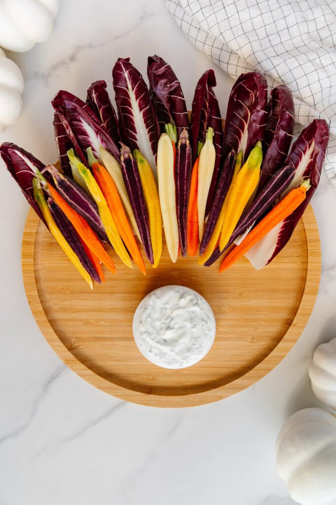 Circular wooden platter with a small bowl of vegan ranch at the bottom and endive leaves and rainbow carrots fanned out at the top.