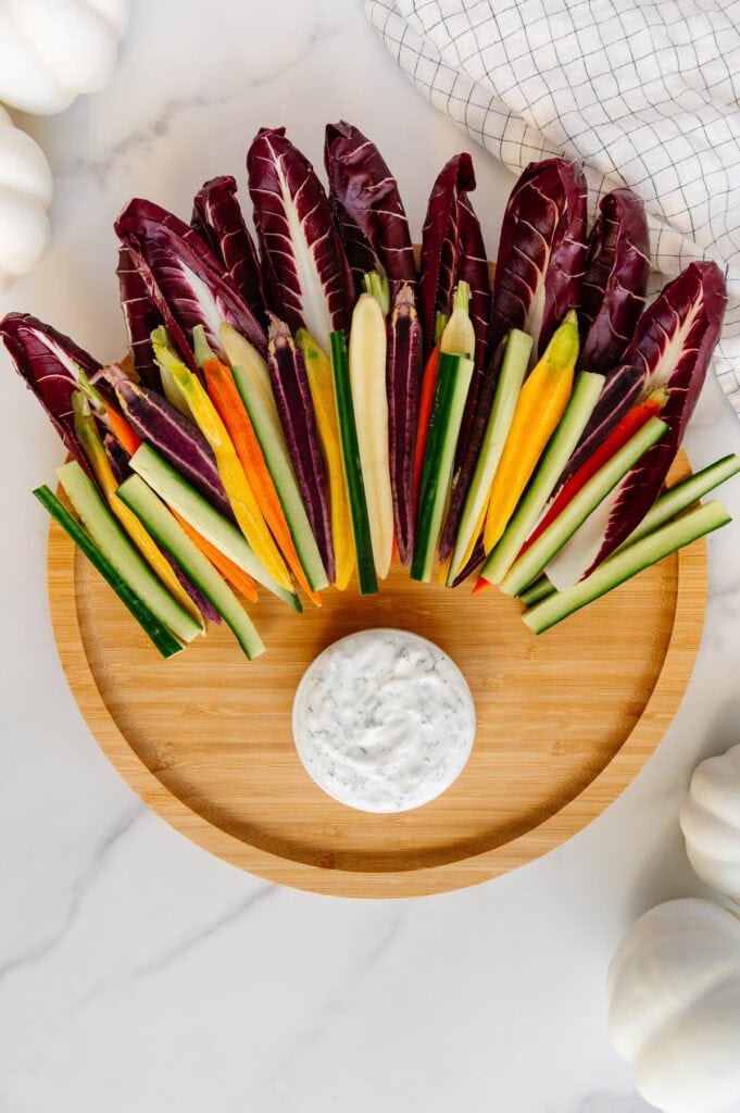 Circular wooden platter with a small bowl of vegan ranch at the bottom and endive leaves, rainbow carrots and Persian cucumbers at the top.