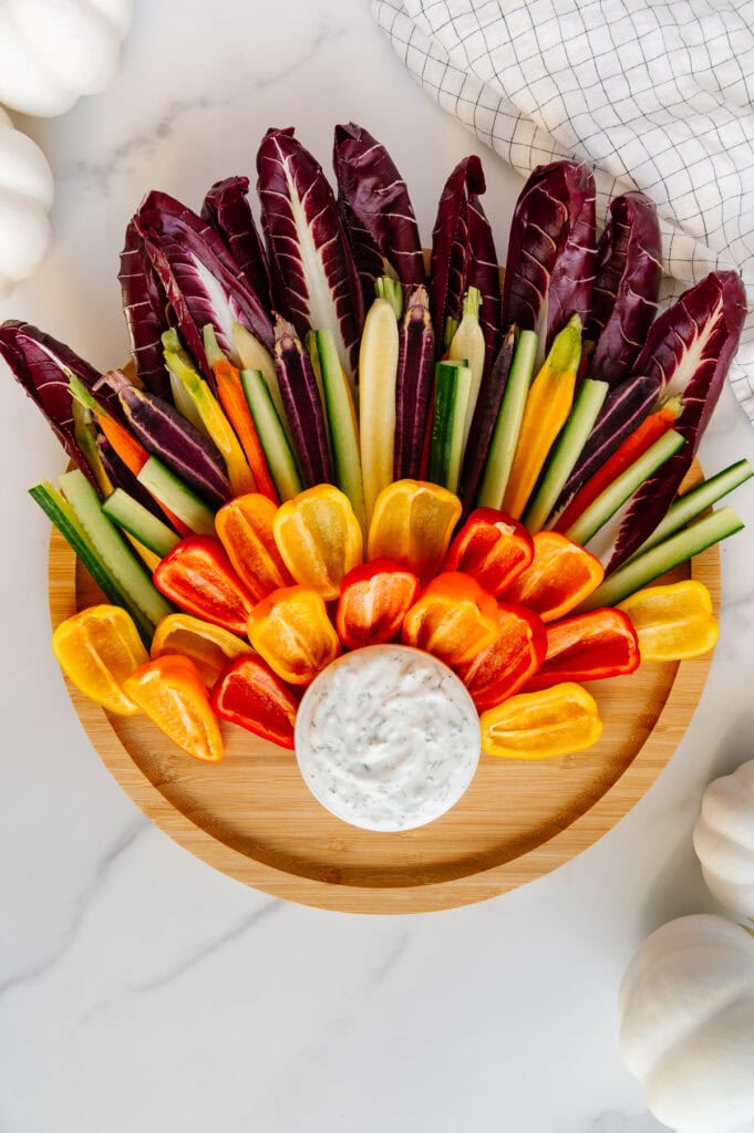 Circular wooden platter with a small bowl of vegan ranch at the bottom and fresh veggies around it shaped like a turkey's feathers.