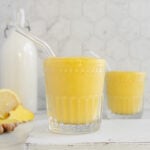 Two glasses of ginger smoothie with glass straws with a bottle of almond milk and sliced lemon and fresh ginger in the background on a white marble surface.
